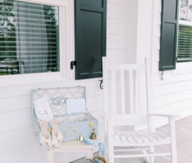 “Bridal details displayed on a white porch with a rocking chair, including blue cowboy boots, bow heels, a light blue suitcase with invitations, and a garter.”