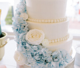 Close-up of a three-tier white wedding cake decorated with white roses and light blue hydrangeas cascading down the side, displayed on an ornate silver cake stand.