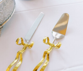Cake knife and server with gold ribbon handles on white tablecloth beside silver cake stand and stacked paper plates.