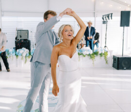 Bride and groom dancing during their wedding reception, smiling as the groom twirls the bride under a white reception tent decorated with string lights and blue floral arrangements.