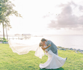 Bride and groom sharing a romantic dip-and-kiss on a grassy waterfront at sunset, with the bride’s long veil flowing behind them and soft light reflecting off the water.