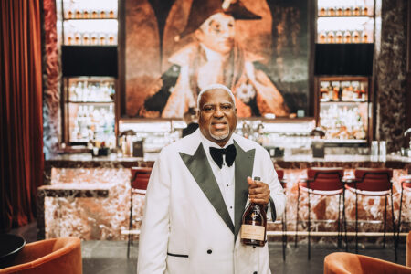 Man in white tuxedo with black trim holds bottle at a bar with red chairs and a large portrait, evoking New Orleans elegance.