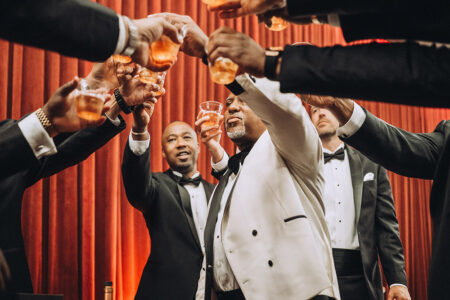 Men in tuxedos toast together before red curtains at a lively New Orleans wedding.