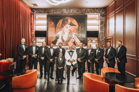 Fifteen in tuxedos pose in an elegant room with red chairs and historic art, inspiring classic New Orleans weddings.