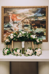 Five white rose bouquets with greenery sit on a white mantel below an ornate painting, perfect for a New Orleans wedding.