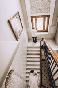 Bride in white dress ascends marble stairs to meet partner by window in elegant, bright New Orleans wedding hallway.