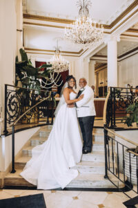 Bride in white gown and man in white tuxedo smile on grand staircase, elegant chandelier-lit Gulf South wedding.