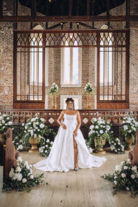 Bride in white gown smiles in a classic New Orleans church with flowers, greenery, stained glass, and brick walls.