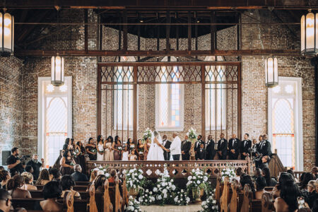 Rustic church wedding with high ceilings, brick walls, and white flowers on pews; bride and groom at altar with wedding party.