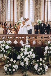 Bride and groom kiss at the altar in a New Orleans church, surrounded by their wedding party, flowers, and watching guests.