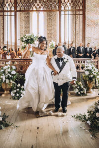 Bride in a white gown and older man in a tux walk joyfully down a flower-lined church aisle, New Orleans wedding.