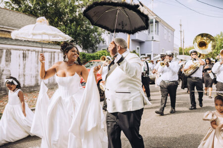 Bride and groom dance with umbrellas in a lively New Orleans wedding parade, followed by brass band and flower girls.