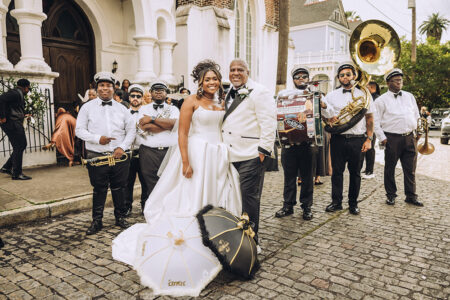 Bride and groom with umbrellas smile before a church, joined by a brass band on a cobblestone street at a Louisiana wedding.