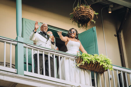 Smiling couple in formal wear waves from a balcony with green shutters and flowers, inspired by New Orleans weddings.