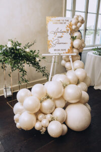 Wedding welcome sign with white and gold balloons and greenery by a window reads: Welcome to the wedding of Antoinette and Sherman, 05 03 2025.
