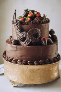 Elegant two-tier chocolate cake with chocolate-dipped strawberries and mask on gold stand for New Orleans or Gulf South wedding.