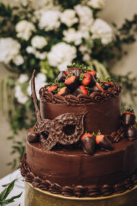 Two-tiered chocolate cake with chocolate strawberries and a mask, set against a floral background—romantic Louisiana wedding inspo.