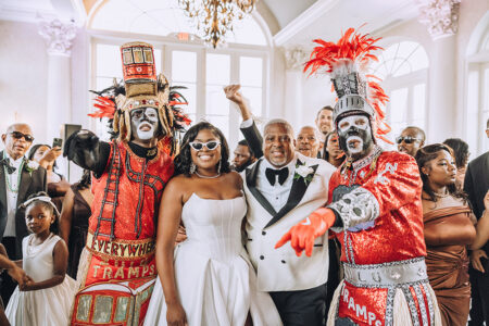 Bride, groom, guests, and Mardi Gras Indian performers in vibrant costumes celebrate at a lively Louisiana indoor wedding.
