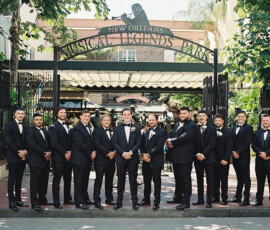 Men in black suits pose and smile in front of the New Orleans Musical Legends Park gate, dressed for a Gulf South wedding.