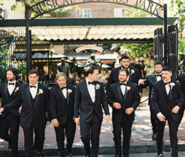 Men in black tuxedos with white boutonnieres stroll beneath a New Orleans Musical Legends Park sign on a sunny day.