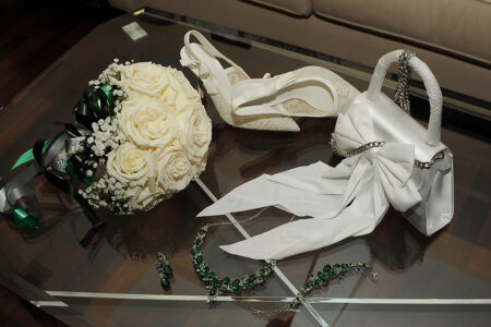 Bridal shoes, white rose bouquet, handbag with bow, jewelry set, and ribbon on a glass table for an elegant Louisiana wedding.