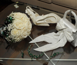 Bridal shoes, white rose bouquet, handbag with bow, jewelry set, and ribbon on a glass table for an elegant Louisiana wedding.