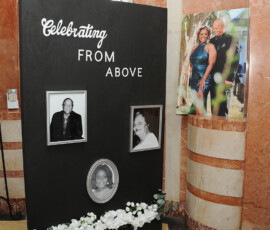 Black memorial board with “Celebrating FROM ABOVE,” black-and-white portraits, and flowers at a Louisiana wedding; smiling couple nearby.