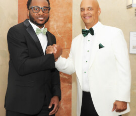 Two men in black and white suits smile and shake hands by a marble wall, reflecting joy and unity—ideal for wedding inspiration.
