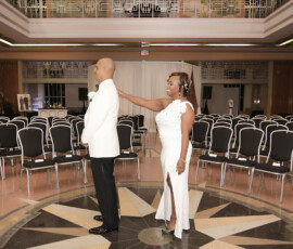 Bride and groom in white attire share a smile in an elegant hall with black chairs—ideal wedding ceremony inspiration.