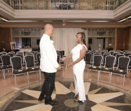 A couple in white formal attire smiles at each other in an elegant Louisiana wedding hall with rows of black chairs behind them.