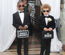 Two boys in tuxedos and sunglasses carry ring cases down a New Orleans wedding aisle, followed by a woman in green.