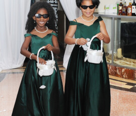 Two young girls in green dresses and sunglasses smile indoors, holding flower baskets with petals at their feet—Gulf South wedding inspiration.
