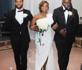 Bride in white dress with bouquet walks between two men in tuxedos down petal-lined aisle at elegant New Orleans wedding.