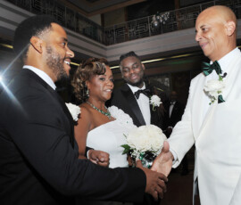 Two men in suits shake hands at a Louisiana wedding, as two women in white dresses with flowers smile nearby.