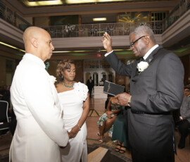 A couple in white faces a suited officiant raising his hand during an elegant ceremony in a grand, sophisticated hall.