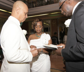 A man and woman exchange rings at an indoor wedding, with an officiant nearby holding a script.