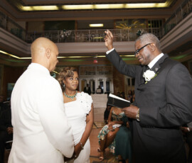 Bride and groom face an officiant raising his hand indoors at a New Orleans wedding, guests seated in the background.
