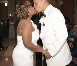 Bride and groom in white kiss and hold hands at their Louisiana wedding as guests watch in the background.