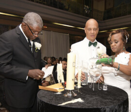 Three at a black table during a New Orleans wedding: one reads, another pours green sand, guests watching in the elegant setting.