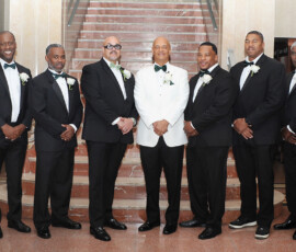 Seven men in tuxedos pose by a marble staircase at a New Orleans wedding; the center wears white with a green bowtie.