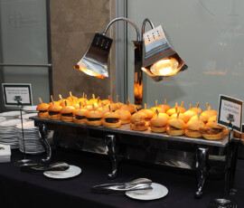 Buffet table with mini cheeseburger and Southern fried chicken sliders under heat lamps, plus plates and utensils.