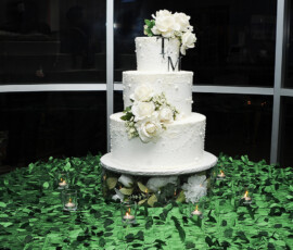 Elegant three-tiered white wedding cake with white roses and initials T & M, on a green leafy tablecloth with candles.