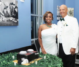 Smiling couple in white formal wear by a decorated cake, leafy tablecloth, and blue walls—elegant New Orleans wedding inspo.