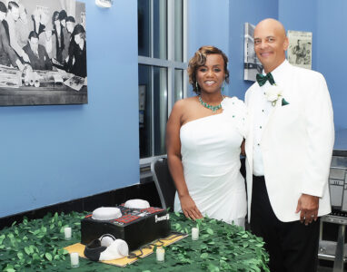 Smiling couple in white formal wear by a decorated cake, leafy tablecloth, and blue walls—elegant New Orleans wedding inspo.