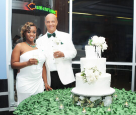 Smiling couple in white attire by a three-tiered wedding cake with TM monogram, holding champagne at a decorated green table.