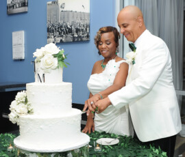 Couple in white smiles as they cut a three-tiered flower cake at their Louisiana wedding, with greenery and photos behind them.