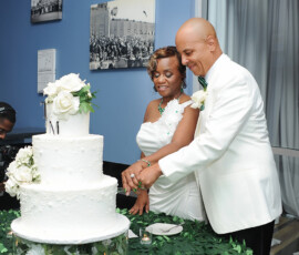 A couple in white formalwear cuts a floral wedding cake at their elegant New Orleans reception, with candles and leaves on the table.