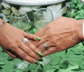 Two hands with wedding rings touch on a leafy green table, with wine glasses in the background for New Orleans wedding inspiration.