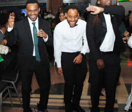 Three men in formalwear laugh and dance at a lively Gulf South wedding, surrounded by guests and joyful celebration.