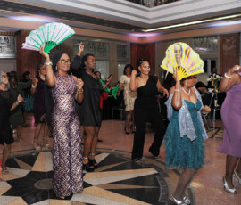 Women in formal dresses dance indoors with colorful fans at a lively, well-lit Louisiana-inspired wedding event.